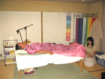 A person lies on a treatment table covered with a pink blanket, while a practitioner sits nearby, preparing for a therapeutic session in a calming space.